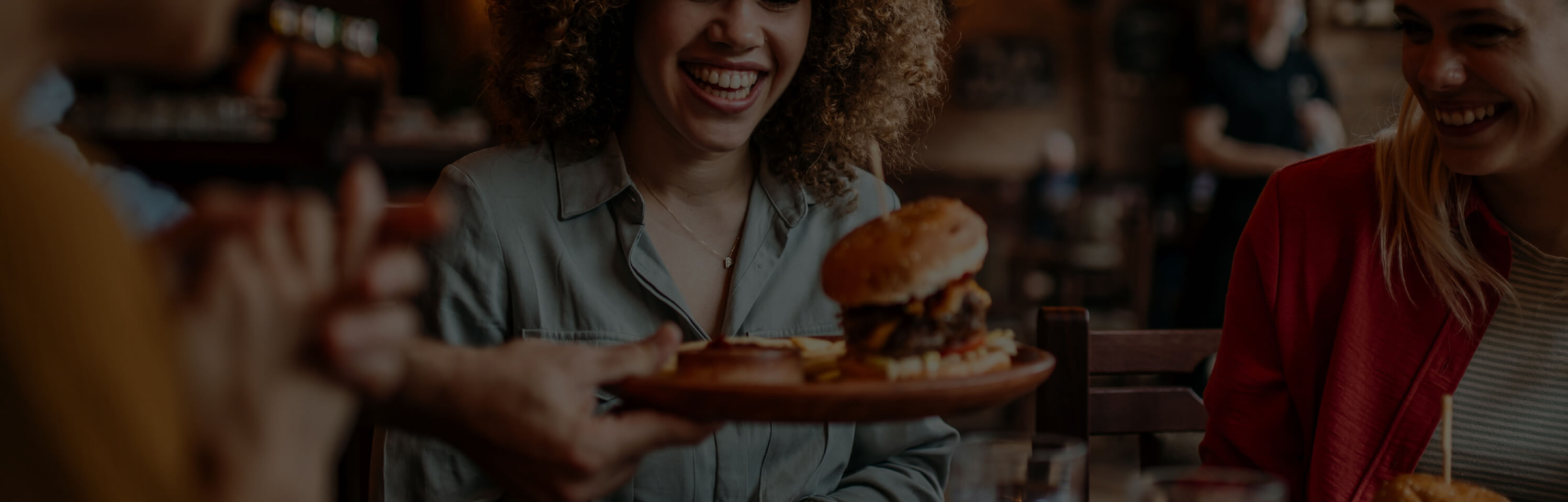 A group of friends sits at a restaurant, smiling and laughing as a server hands a plate with a burger and fries to a woman with curly hair. The atmosphere is warm and lively, and the table is set with drinks and other dishes.