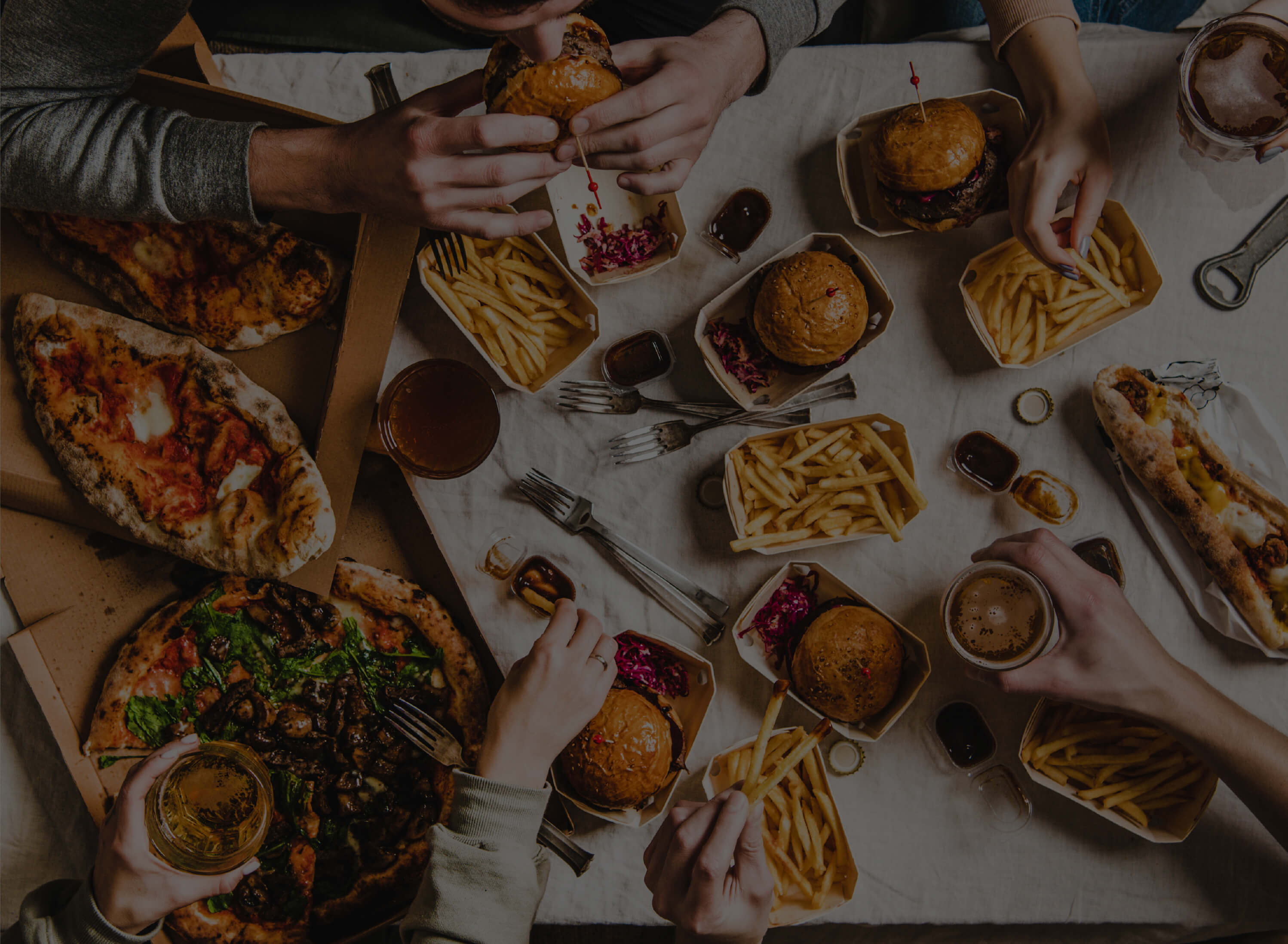 A group of people eating various foods including burgers, fries, and pizzas on a table. Multiple hands are reaching for the food and holding drinks. Table setting includes cutlery and cardboard boxes.