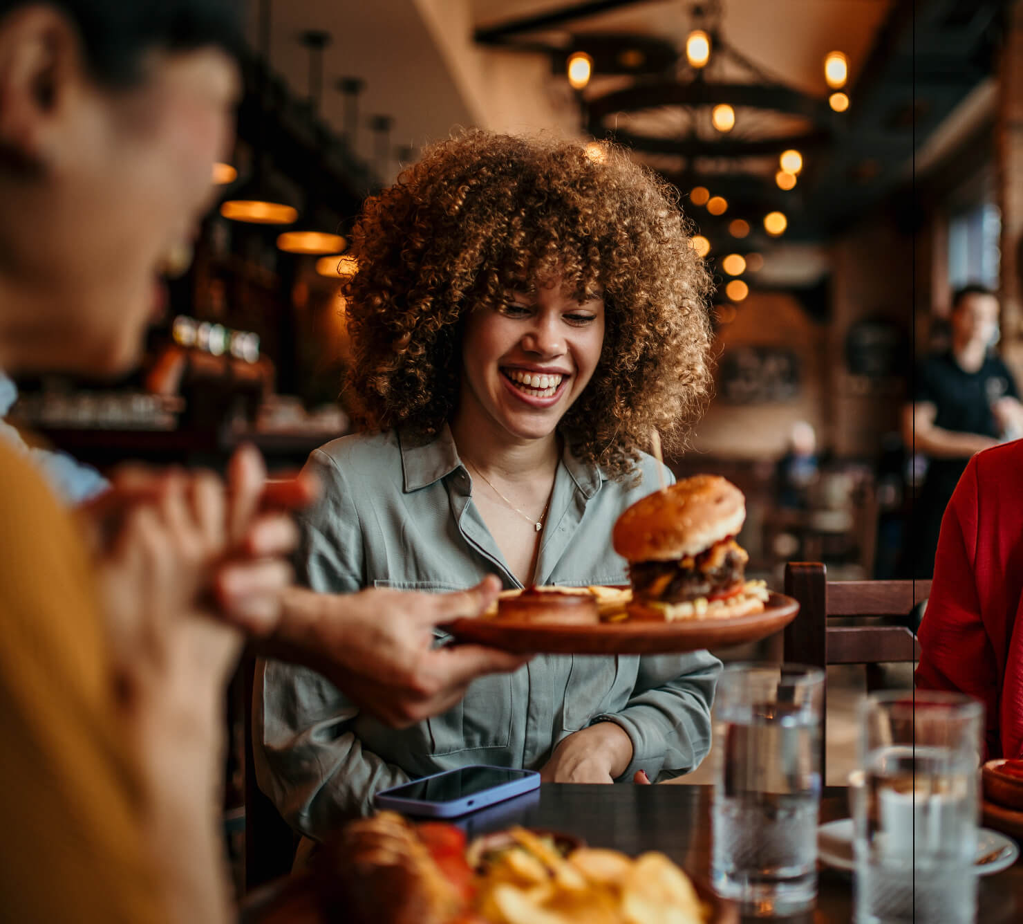 A woman with curly hair smiles as a server places a burger in front of her at a restaurant. Two other people are partially visible, and the setting is warmly lit with hanging lamps.